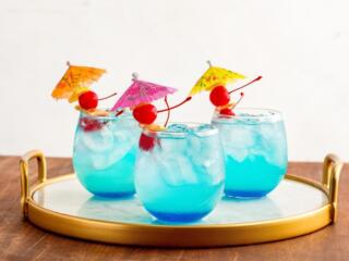 3 glasses of blue mermaid lemonade cocktails with cherries and mini umbrella garnishes