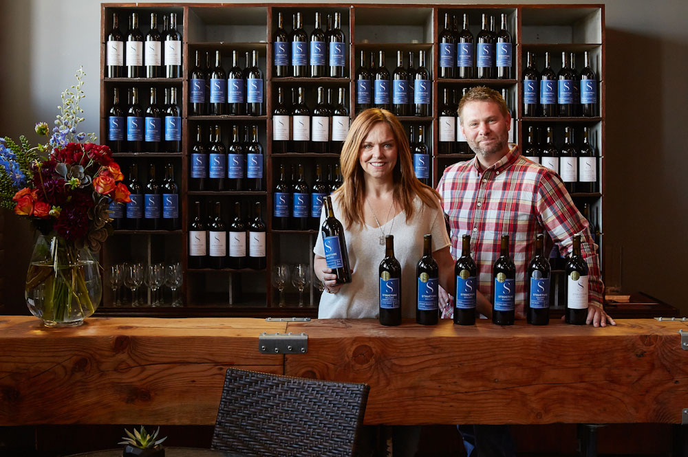 Two winemakers smiling and standing in front of a wall of locally-made wine. 
