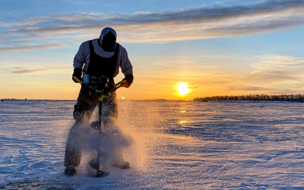 Man drilling a hole into the ice with an auger and pictured in front of a gorgeous sunset. 