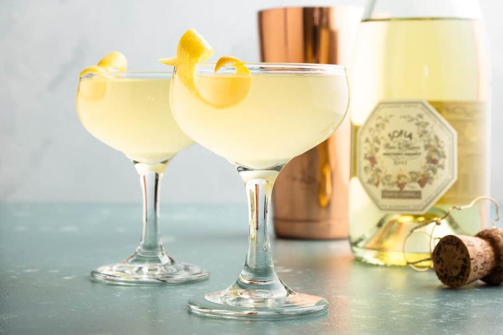 French 77 cocktails topped with a lemon twist situated beside Champagne and a cocktail shaker. 