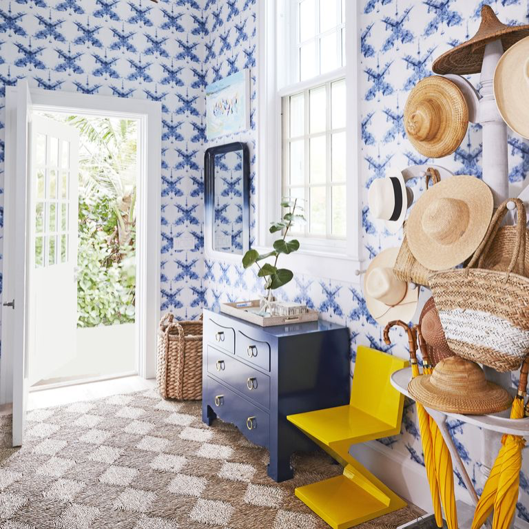 entry foyer with canary yellow zig-zag stool, umbrellas, blue patterned wallpaper, hat collection, woven handbags, woven patterned rug, blue painted chest of drawers