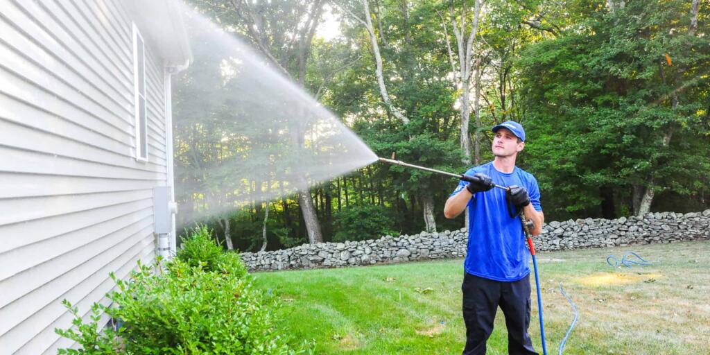 guy in blue shirt pressure washing the side of a house, stone wall in background