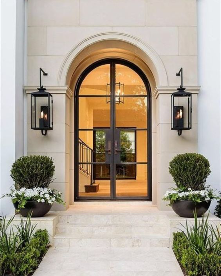front entrance greenery, potted flowers and potted plants, white stone walkway, arched glass door with metal accents, black metal lighting sconces flanking door
