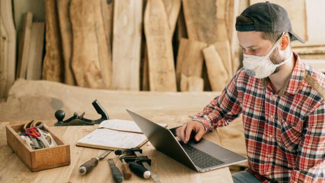 man in a workshop with wood, tools, a book, and a laptop