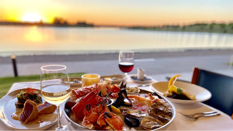 plates of seafood and fish with glasses of wine overlooking lake at sunset