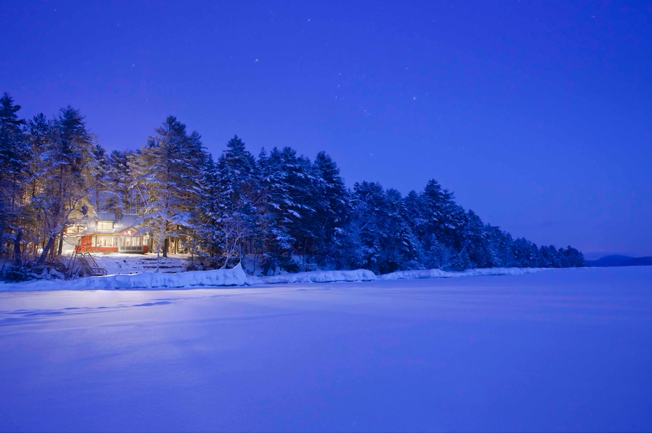 exterior of home lit up at night on the shoreline of a snow-covered Lake Winnipesaukee in New Hampshire