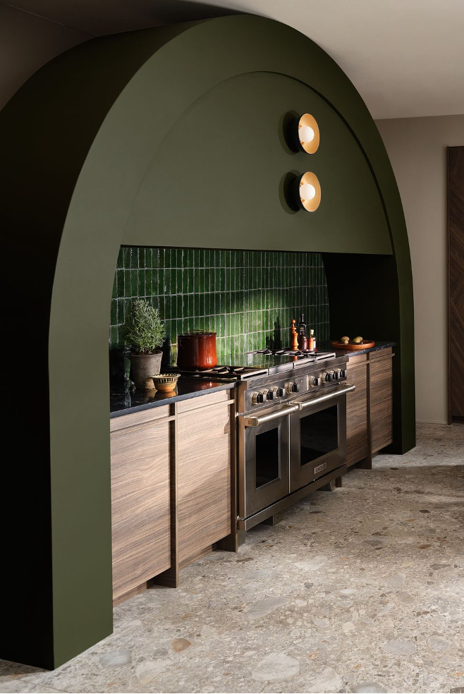 Statement range hood in dark olive-green with accent lights and a green tile backsplah, stone flooring