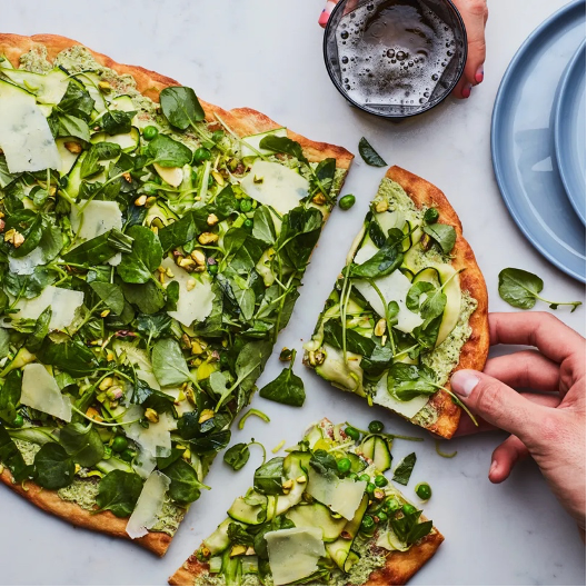 White pizza with shaved vegetables and pesto with plates, fingers, fingers holding beverage