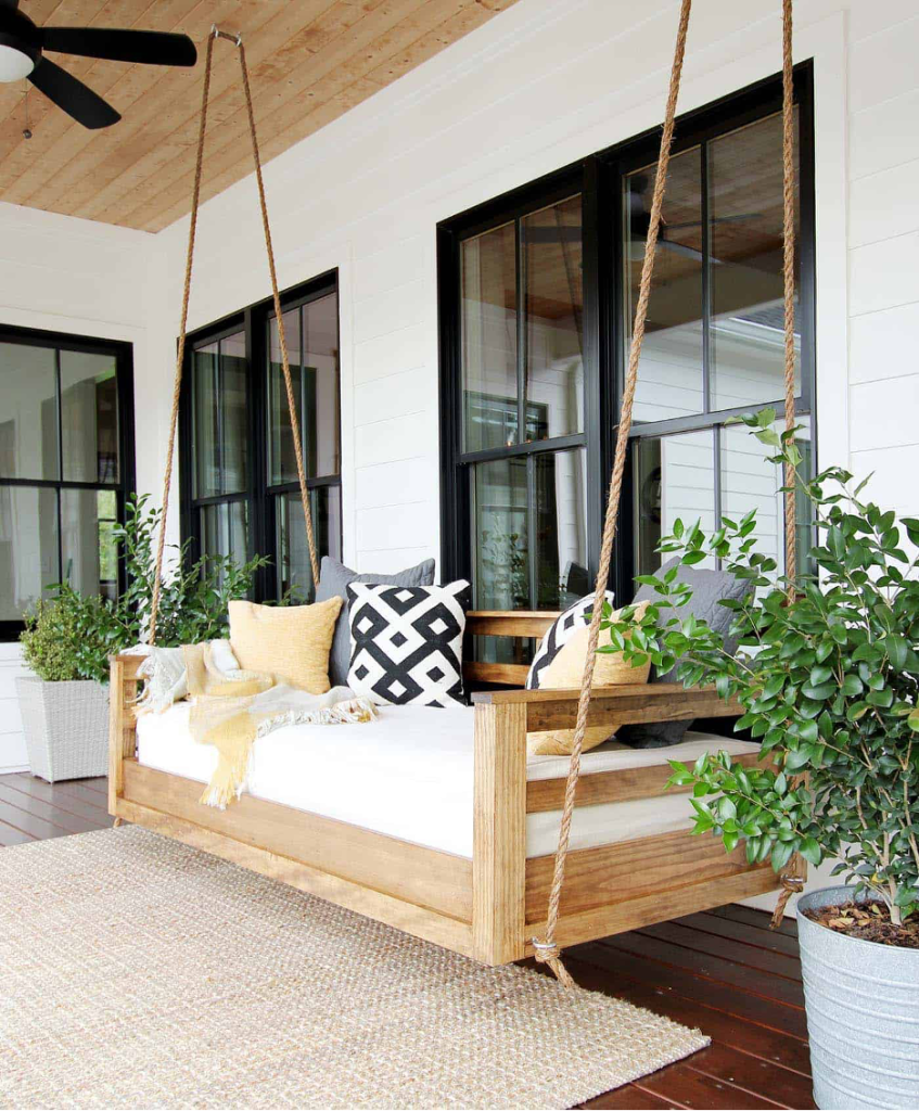 Natural wood swing bed on front porch with potted plants, woven rug, and black trimmed windows