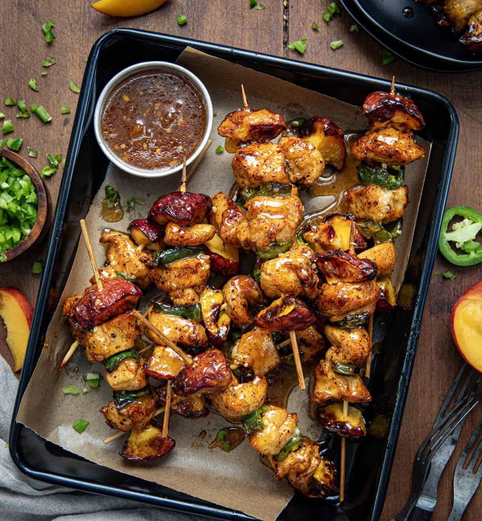 Sweet Heat Chicken Skewers with Peach Glaze in a roasting pan woth a side of peach glaze in the pan served on rustic wooden table with jalapeno, peach, onion garnishes on the side.