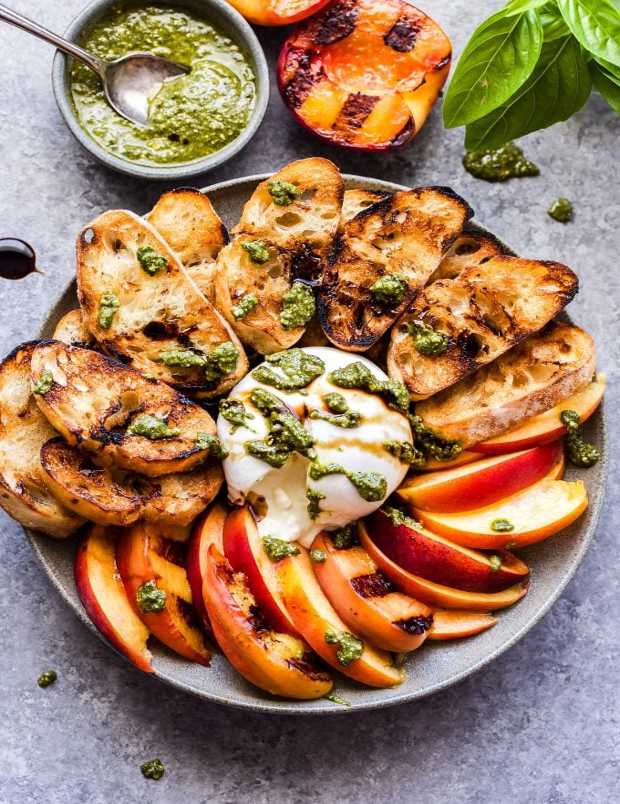 Grilled Peach-and-Burrata Crostini served on platter with pesto and a grilled peach half and fresh basil in background