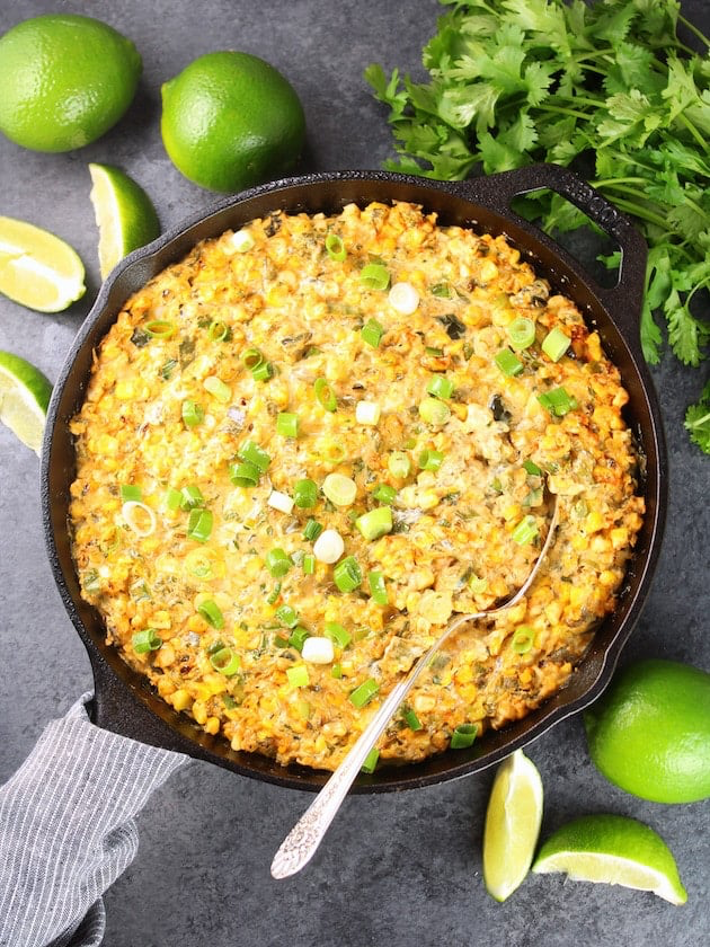 Charred Poblano Corn Dip in iron skillet with limes and herbs in background