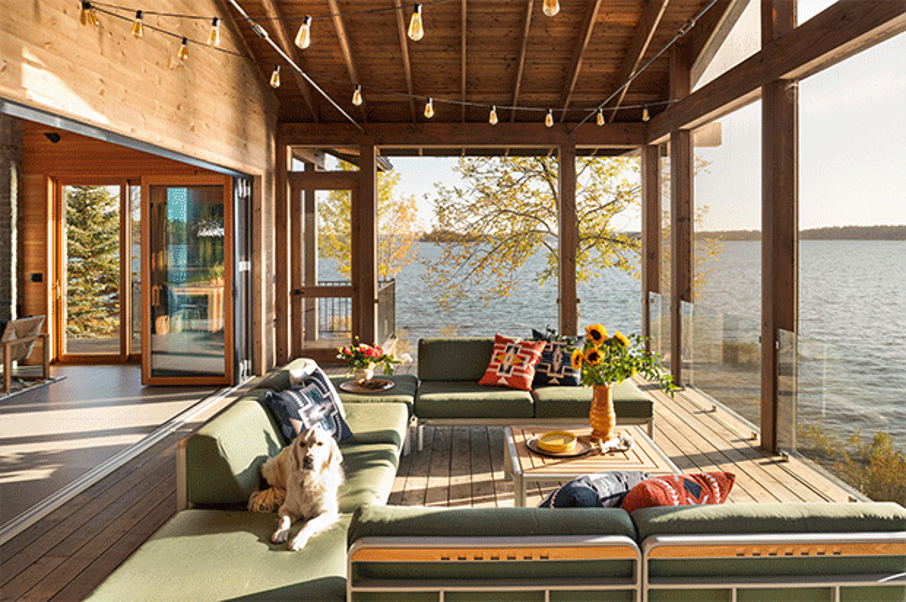 Covered deck overlooking Lake Huron, incorporating plexiglass deck railing, modular seating, wooden-topped coffee table with metal trim, colorful pillows and accessories, sliding glass door connecting to interior, string lights overhead, family dog lounging on the outdoor sofa, 