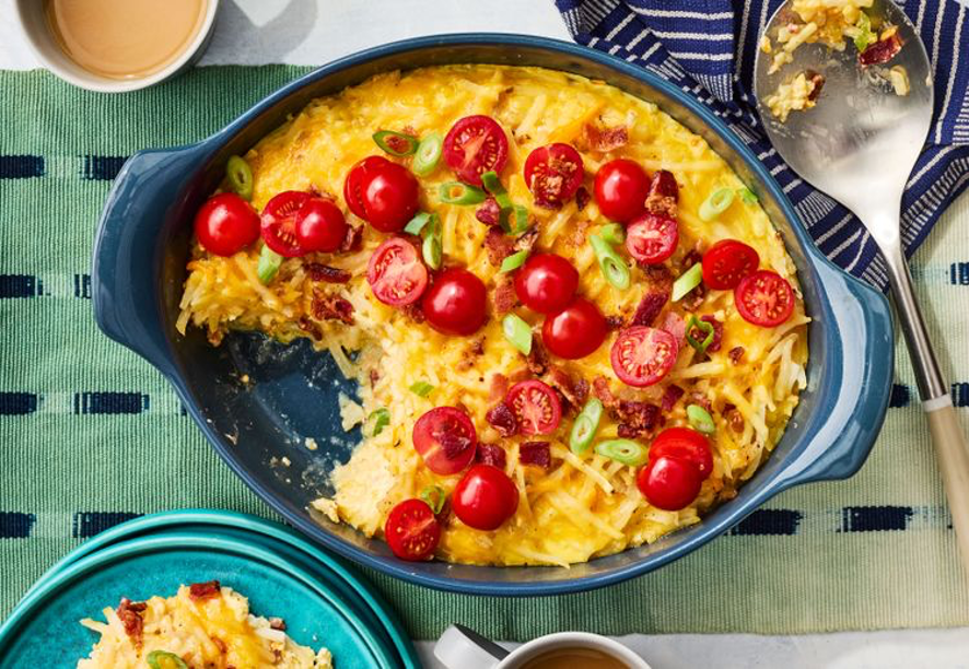 Deep-Dish Loaded Hash Brown Casserole topped with cherry tomatoes in a blue baking dish on a green and blue napkin with other plates, cups, and serving spoon in background