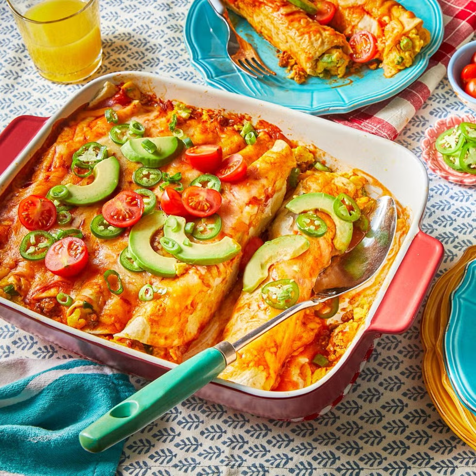 Breakfast Enchiladas in a casserole dish topped with avocados, jalapenos, and halved cherry tomatoes with serving spoon