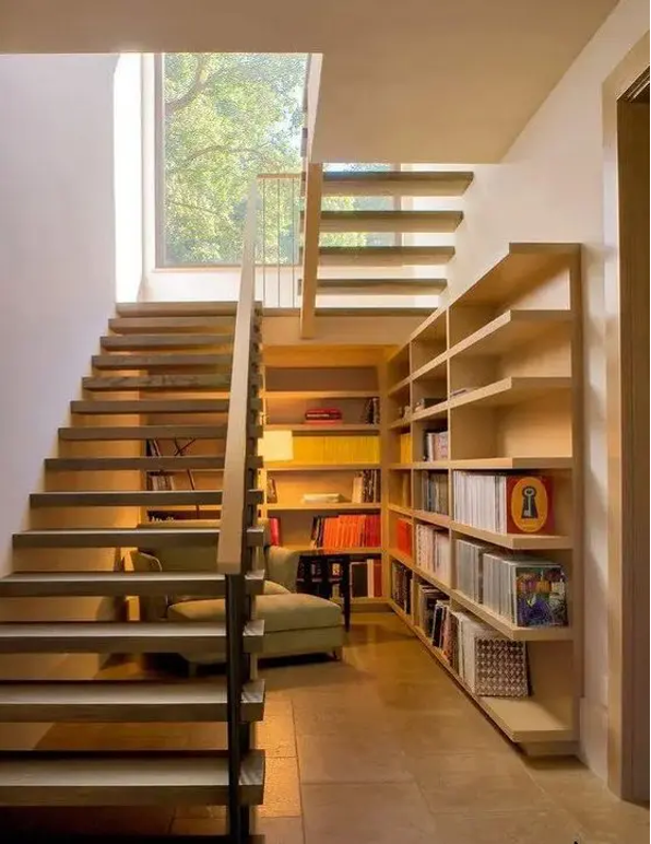 Tucked-away reading nook underneath an open staircase with book shelves and a lounge chair; contemporary design with landing featuring a picture window looking out onto the treetops