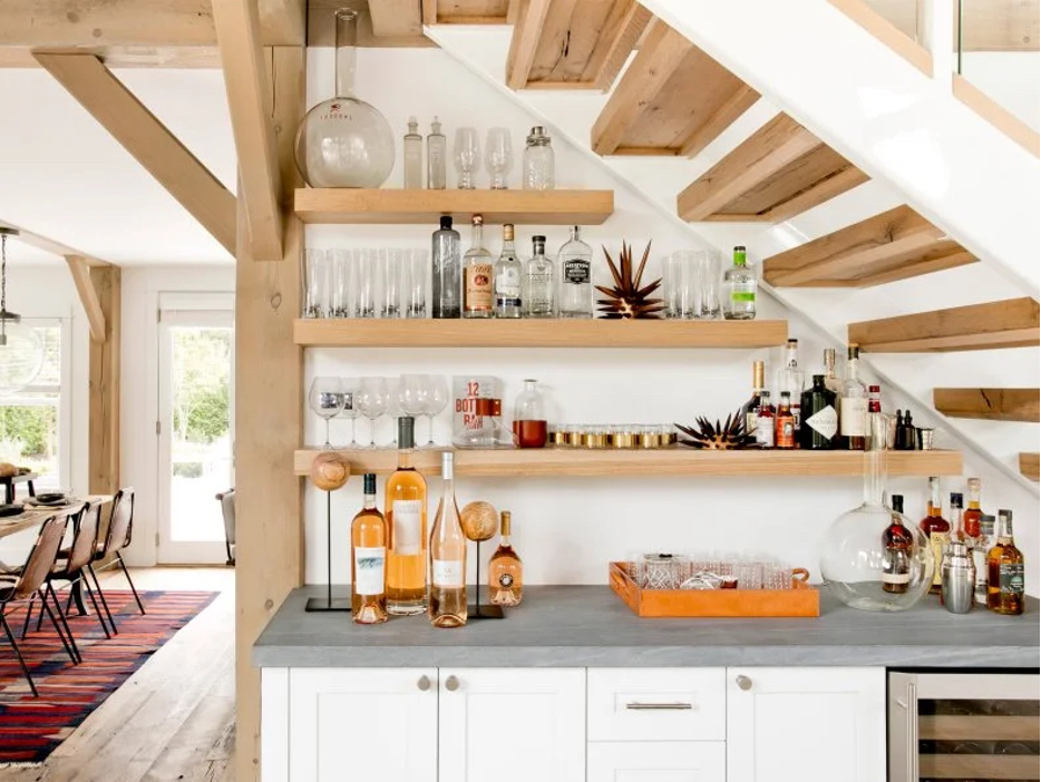 A bar area tucked underneath the stairs, with open wood shelving, stone countertop, refrigeration, and cabinetry; modern design
