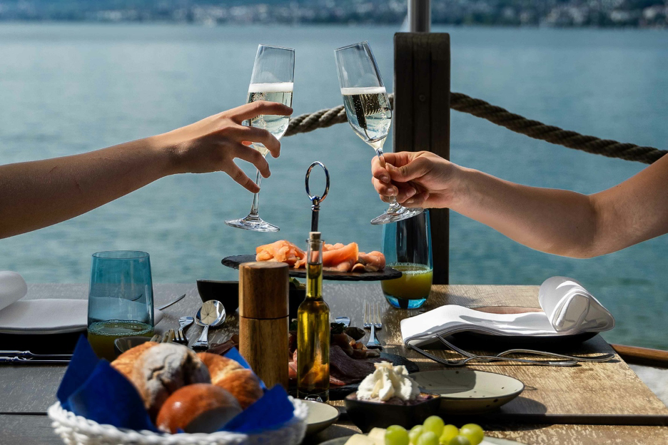 arms and hands toasting champagne glasses across a table while on a dock setting with brunch fare overlooking the blue lake waters
