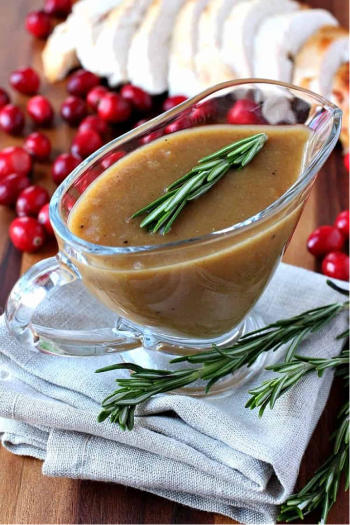 Bourbon Cranberry Turkey Gravy in a glass gravy boat and garnished with rosemary sprig and cranberries and sliced turkey in the background